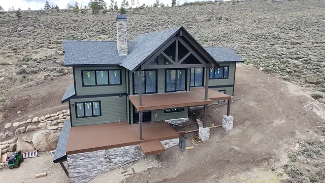 Aerial view of a green board-and-batten custom home with timber-frame gable, stone chimney, and wraparound composite decks, built by Rondeau Construction in Grand County, Colorado